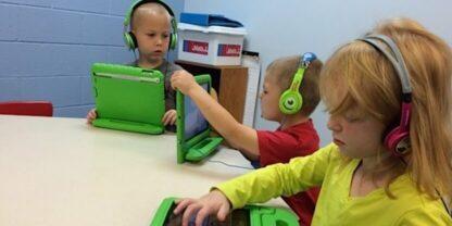 Three students wearing headphones and working on tablets at a large round table.