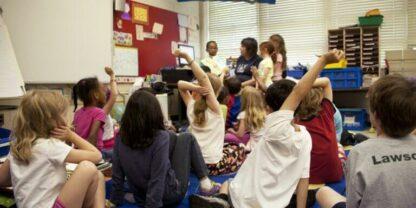Students sitting on the classroom floor with some raising their hands.